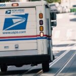 A USPS mail delivery truck driving uphill in a city neighborhood on a sunny day, with a view of buildings and trees in the background.