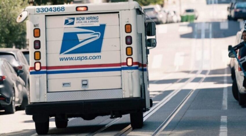 A USPS mail delivery truck driving uphill in a city neighborhood on a sunny day, with a view of buildings and trees in the background.