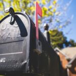 Close-up of a black mailbox with a raised red flag, set against a sunny suburban backdrop with trees and a house in the distance.