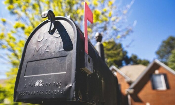 Close-up of a black mailbox with a raised red flag, set against a sunny suburban backdrop with trees and a house in the distance.