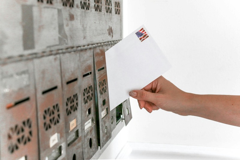 A person placing a white envelope with an American flag stamp into an old, metal mailbox, symbolizing political mail and election-related correspondence.